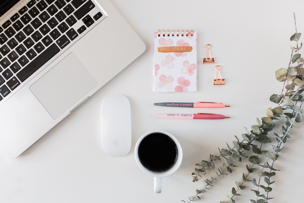 Image of a desk with a laptop, notebook, pens, and coffee on it.